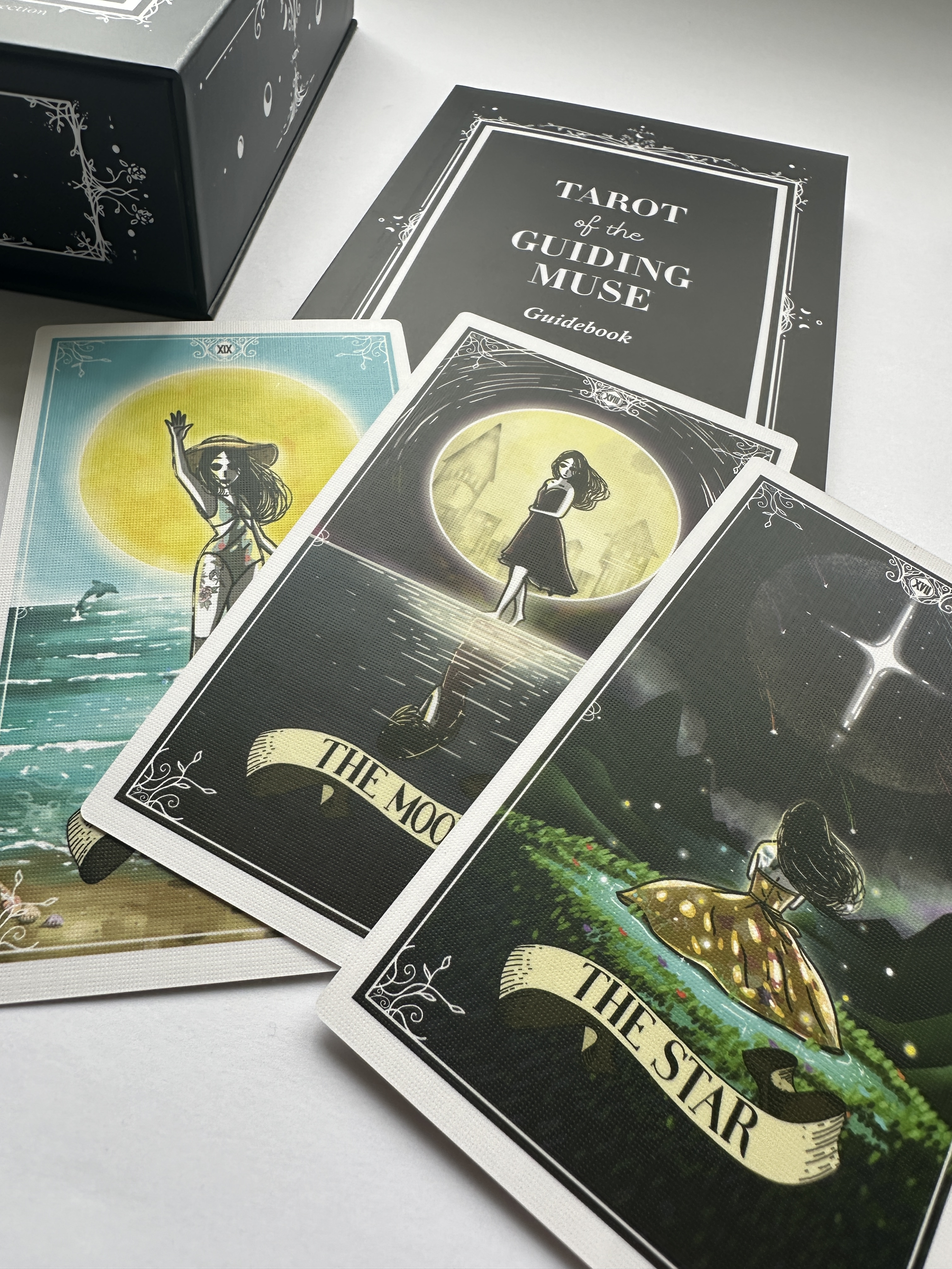 Tarot cards from 'Tarot of the Guiding Muse' set on a white surface with a box in the background.